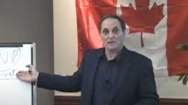 A man in a suit with a Canadian flag behind him points to a whiteboard he has written on.