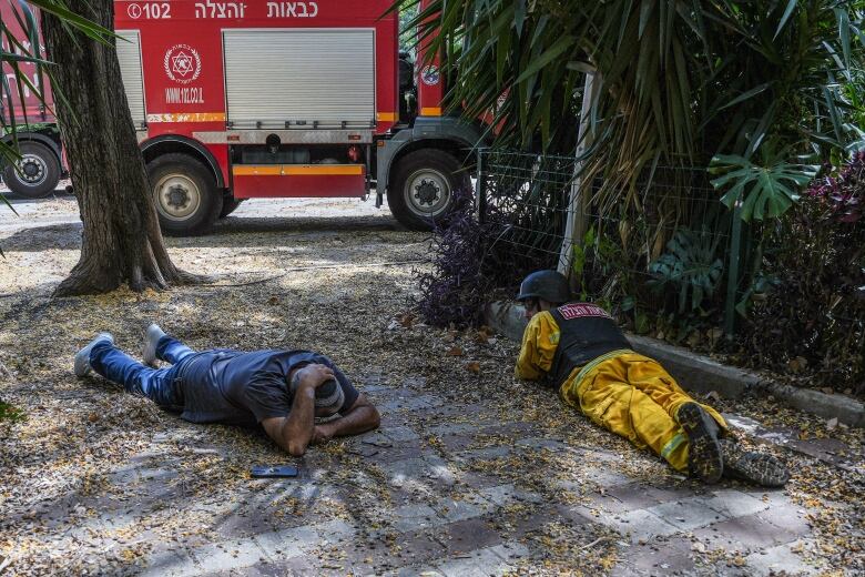 Two people who appear to be men, one wearing a helmet lie on the ground covering themselves near a fire truck.