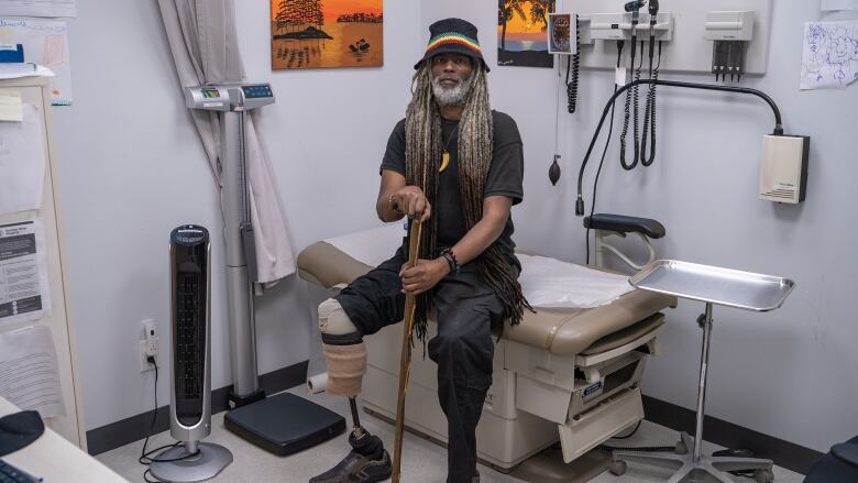 Man wearing a hat sits on an examination table in a clinic with a cane.