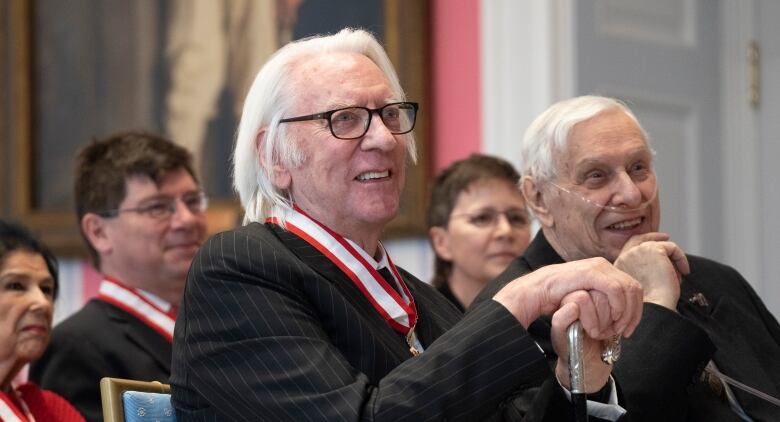 A grey-haired man in a dark suit sits with an ornate medal around his neck at a ceremony.