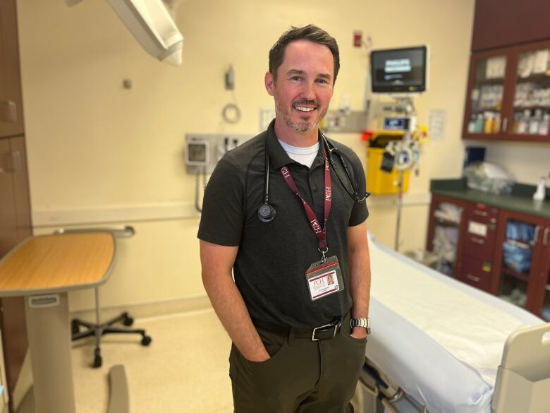 A doctor stands in the middle of a treatment room with a bed and monitors behind him. 