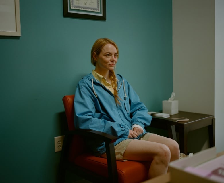 Actress Emma Stone is seen sitting in a chair in front of a blue wall in the movie Kinds of Kindness.