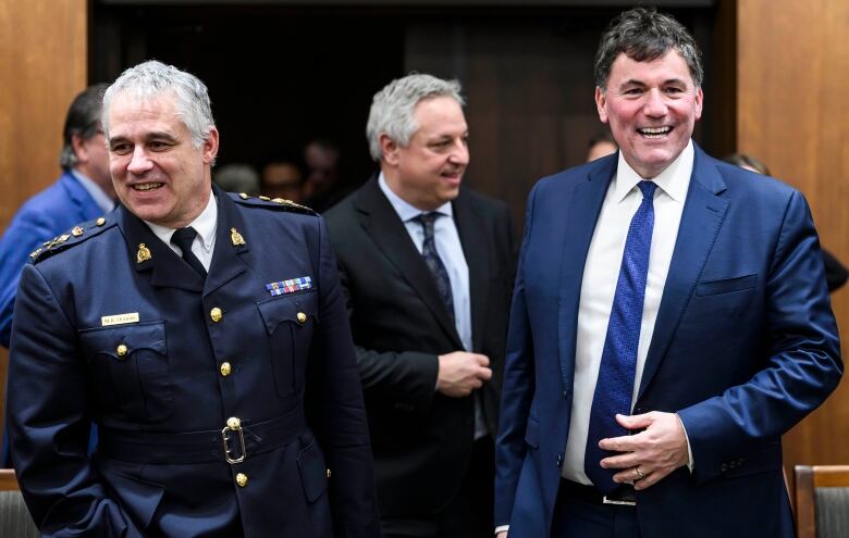 An RCMP head in blue uniform, left, standing, smiling, next to men in business suits, the head of CSIS and the public safety minister