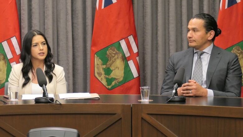 A woman and a man sit before microphones in front of several Manitoba flags.