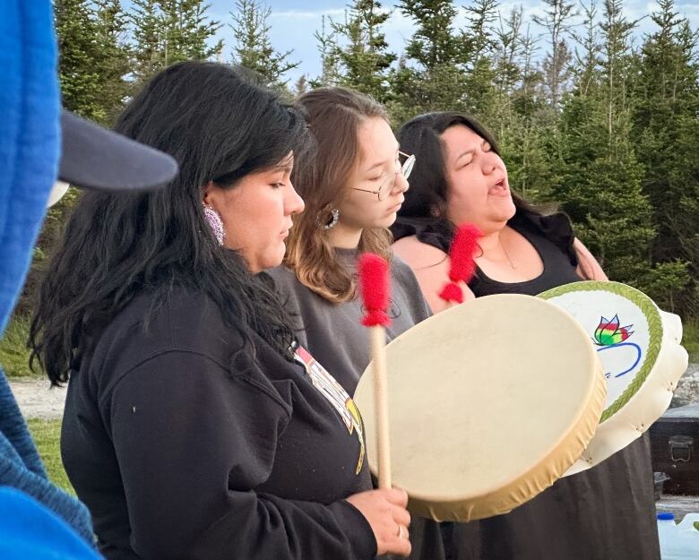 Two young Indigenous women use red mallets to strike Mi'kmaw hand drums while a third woman sings.