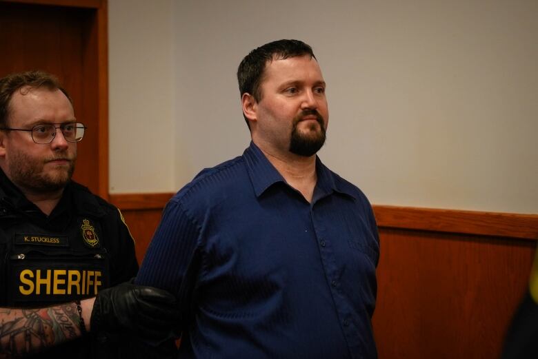 A heavy-set man with a blue dress shirt, dark hair and dark goatee beard is led into a room by a short sheriff's officer with glasses.