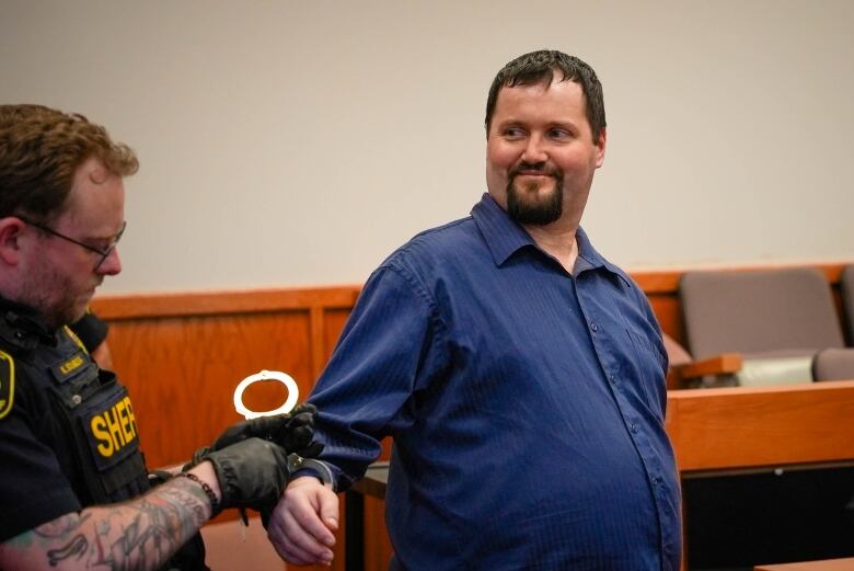 A heavy-set man with a blue dress shirt, dark hair and dark goatee beard looks to the left of the frame while his right hand is being uncuffed.