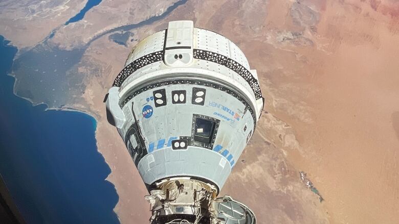 Earth is in the background with the ocean and desert, and in the foreground is a white spacecraft attached to the International Space Station.