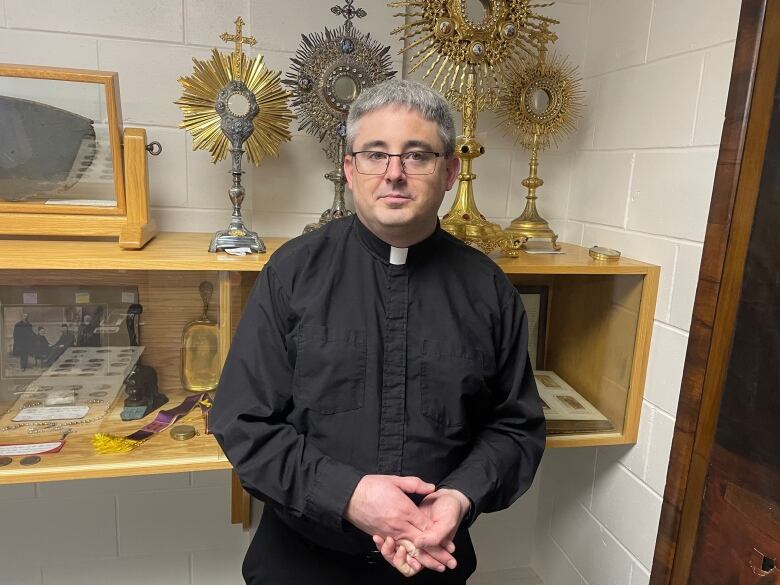 A priest stands in front of a shelf holding old artifacts 