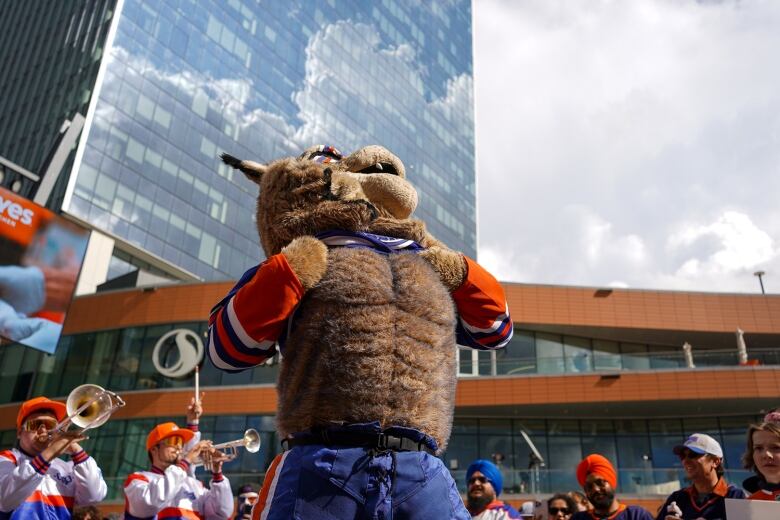 A person in a giant lynx costume stands in a crowd of orange and blue people playing brass instruments.  