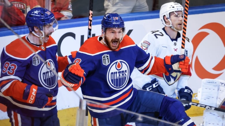 A male ice hockey player pumps his fists while shouting in celebration next to a teammate as an opposing player skates away.