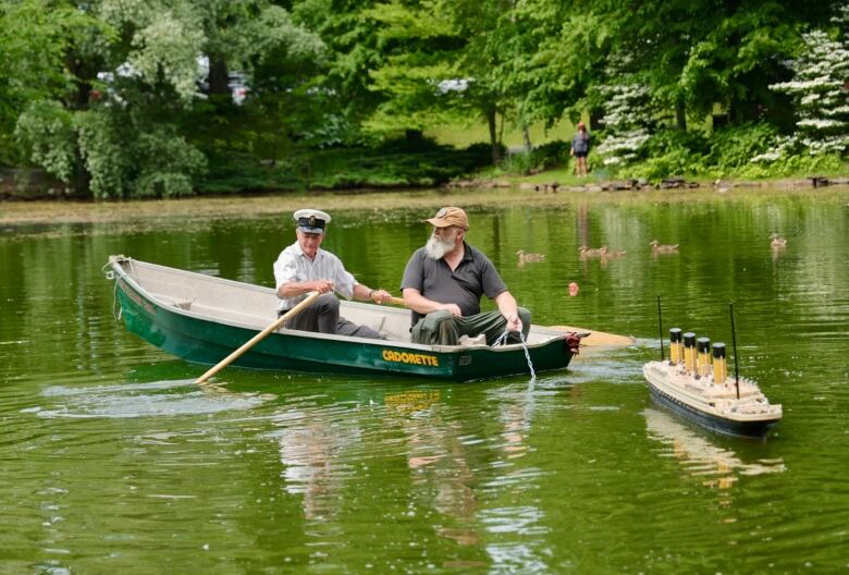 Two men are seen in a rowboat pulling a model of the Titanic behind them