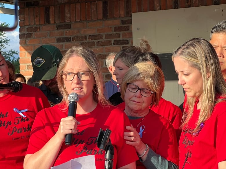 A woman with blonde hair wearing glasses and a red T-shirt that says Light Up the Park holds a microphone and looks emotional