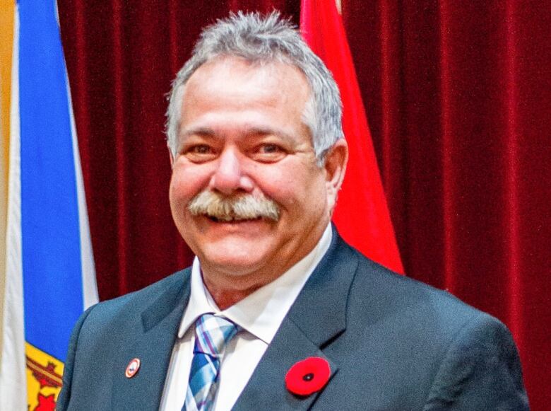 A man with grey hair and moustache wearing a blue suit, white shirt and blue tie smiles in front of a red background with flags in the back.