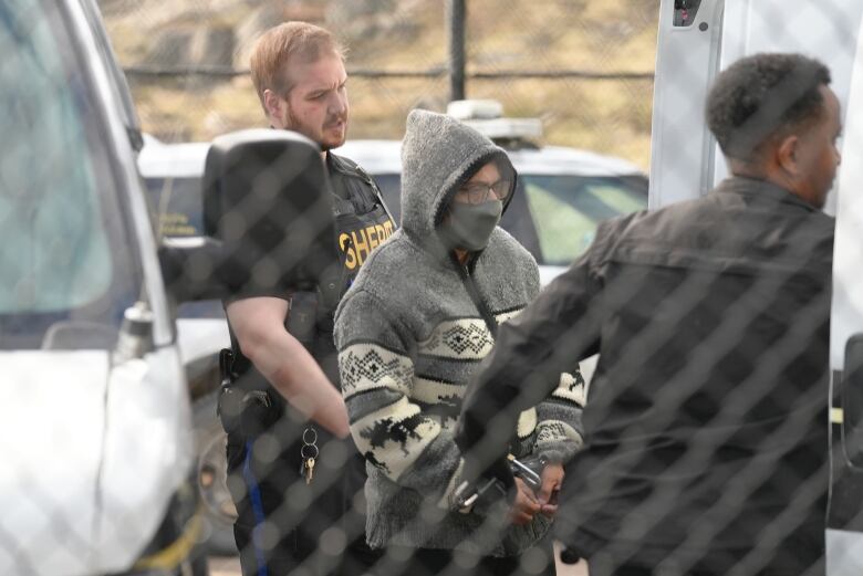 A woman wearing a grey sweater with the hood up and a grey face mask and hand cuffs is seen through a fence with two sheriffs on either side of her. 