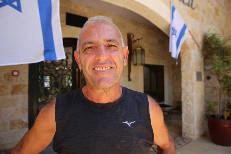 A man with close-cropped white hair wears a black sleeveless shirt while sitting in a stone-walled courtyard draped with Israeli flags. 