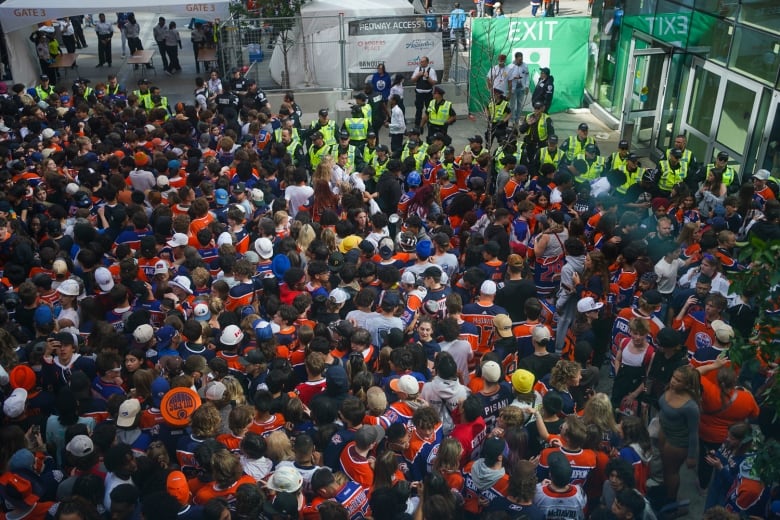 A mass of people wearing blue-and-orange are packed in front of police officers wearing reflective yellow vests, outside of an arena.