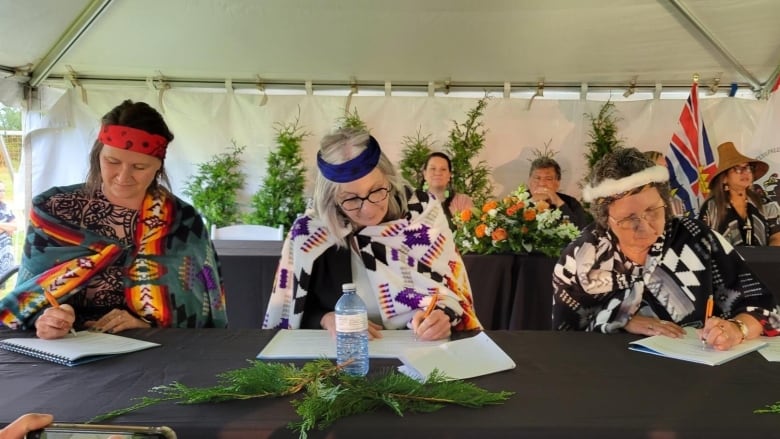 Three women sign an agreement at a table, all of whom are wearing First Nations regalia.