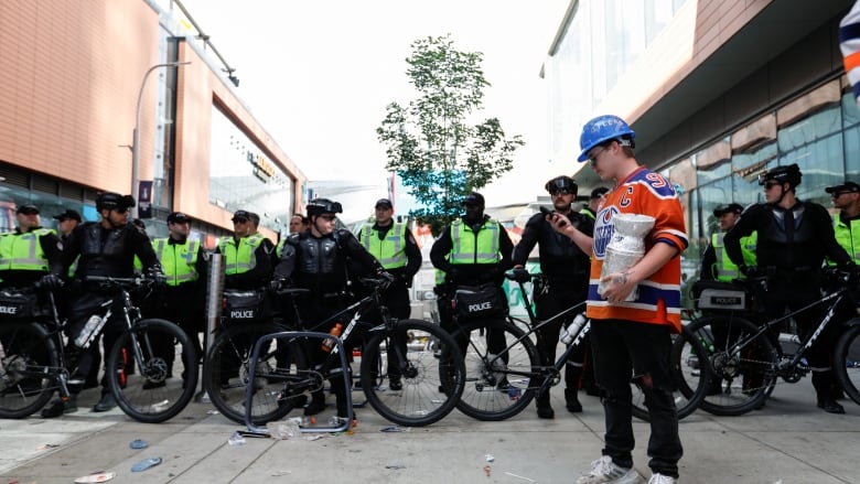 A line of police in neon reflector vests stand behind bicycles behind a man in an orange jersey holding a phone and a large silver cup. 