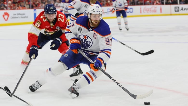 A male ice hockey player controls the puck with both hands on his stick as a defender reaches out with their stick.