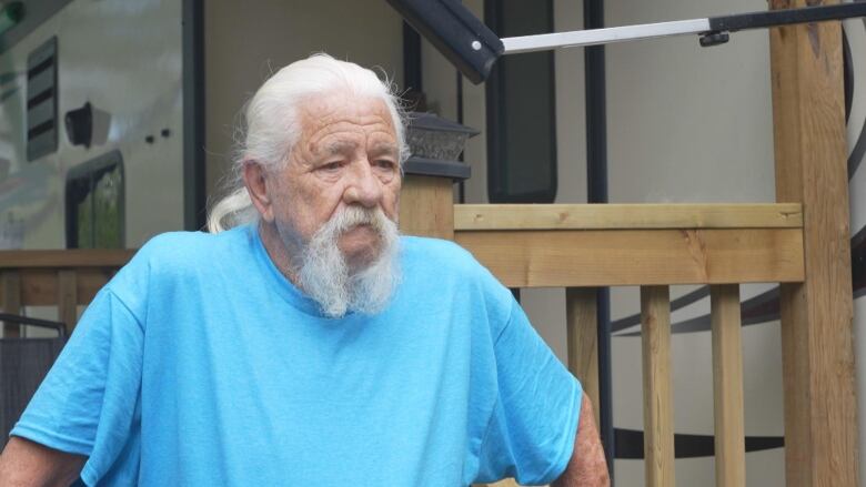 An older man with white hair and a blue T-shirt speaks outside an RV.