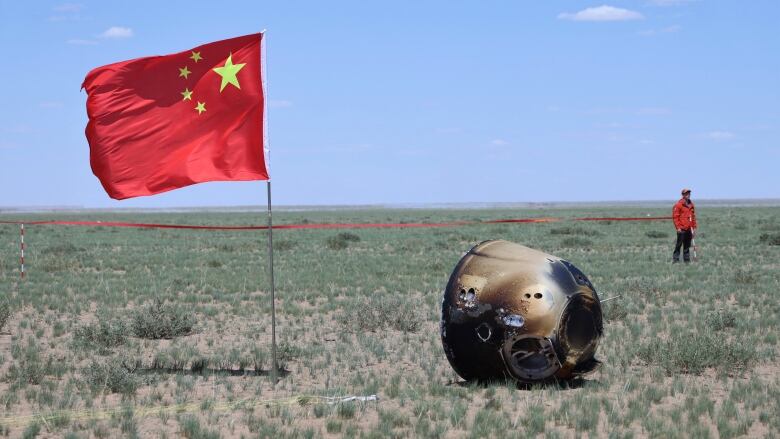 A gumdrop-looking metal probe lies in a field with the Chinese flag flying nearby and a man in a red jacket and red baseball cap in the distance.