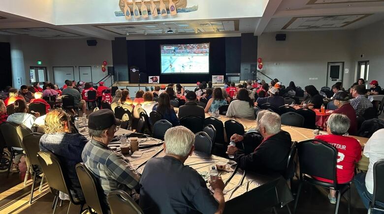 A room of people in a hall sit at tables and watch a hockey game on a screen.