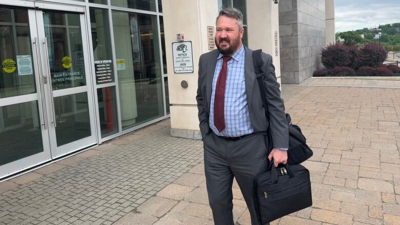 Bearded man in a dark suit and carrying a briefcase walks away from a stone building. 