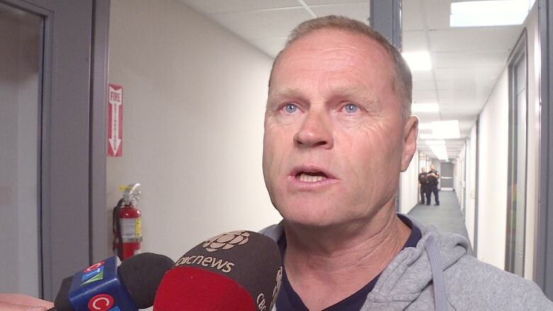 A man with a grey hoodie speaks into a microphone while two police officers can be seen far away down a hallway behind the man.