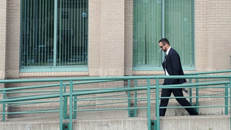 Seen from a distance, a bearded, dark-haired man in a suit looks down as he walks down a ramp from the Prince George courthouse, where he is on trial for obstruction of justice. 