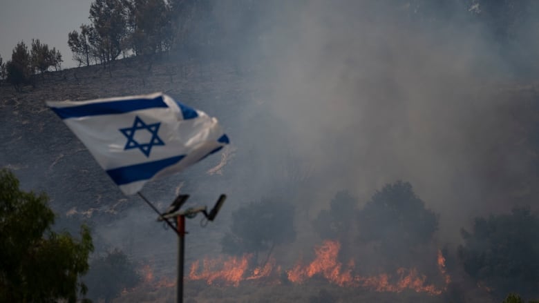 An Israeli flag flutters next to a fire burning in an area near the border with Lebanon in Safed, northern Israel on Wednesday, June 12, 2024. Scores of rockets were fired from Lebanon toward northern Israel on Wednesday morning, hours after Israeli airstrikes killed four officials from the militant Hezbollah group, including a senior military commander.