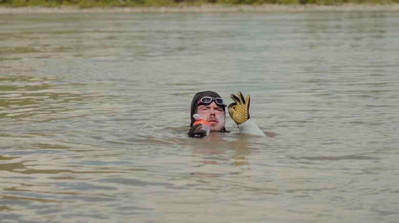 A man in a wetsuit and goggle gives the 'OK' sign from some murky water.