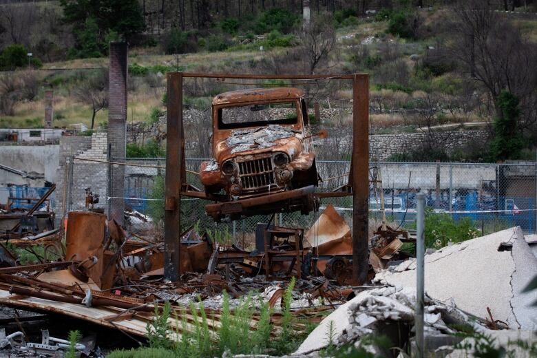 A burned-out car lies suspended on a platform, surrounded by debris and blackened ground.