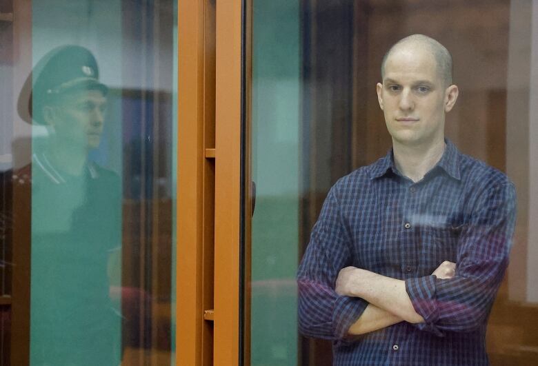 A man in a blue shirt stands with his arms folded inside a glass enclosure.