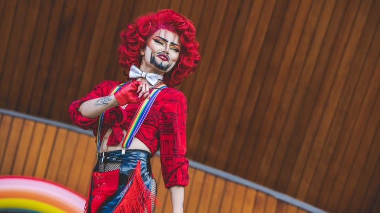 A drag king wears a short, red, curly wig and sports a beard drawn with makeup and a red shirt with rainbow suspenders. 