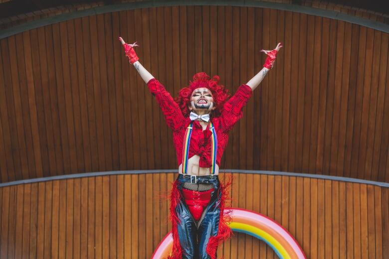 A drag king stands on a stage with his arms thrown up and his head back. He's wearing a short, curly red wig, a red cropped blouse, and leather chaps.