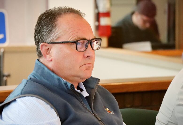 A man with black framed glasses wearing a light blue shirt and a dark blue vest is sitting.