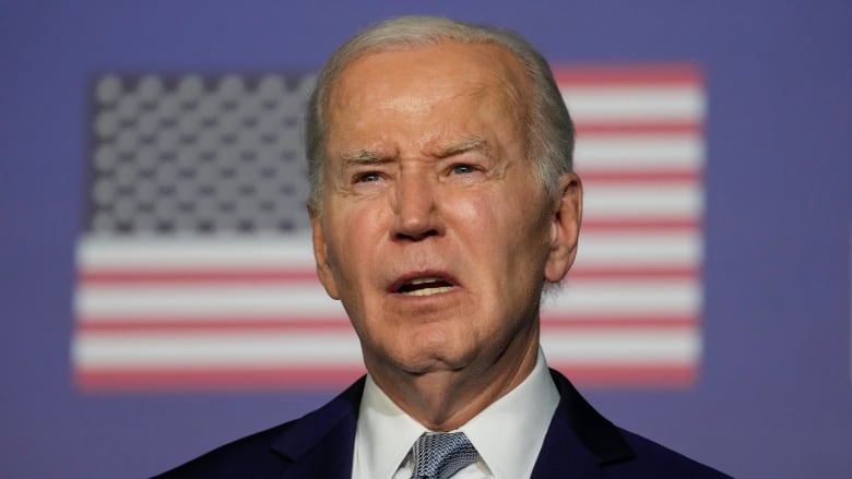 A man in a dark suit with a blue tie and an American flag pin on his lapel stands at a podium in front of a large American flag.