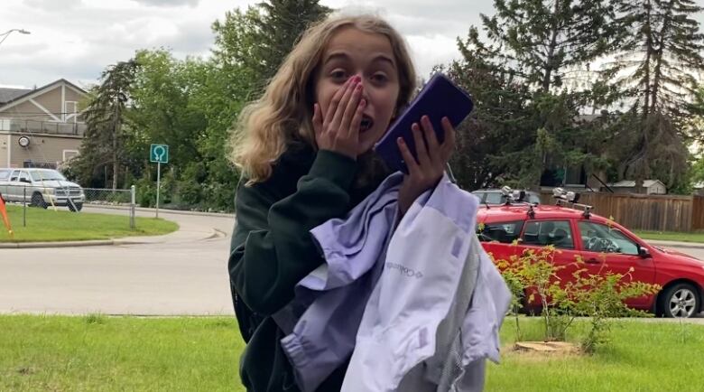 A young girl with long blonde hair covers her mouth while holding a cell phone and standing in a yard.