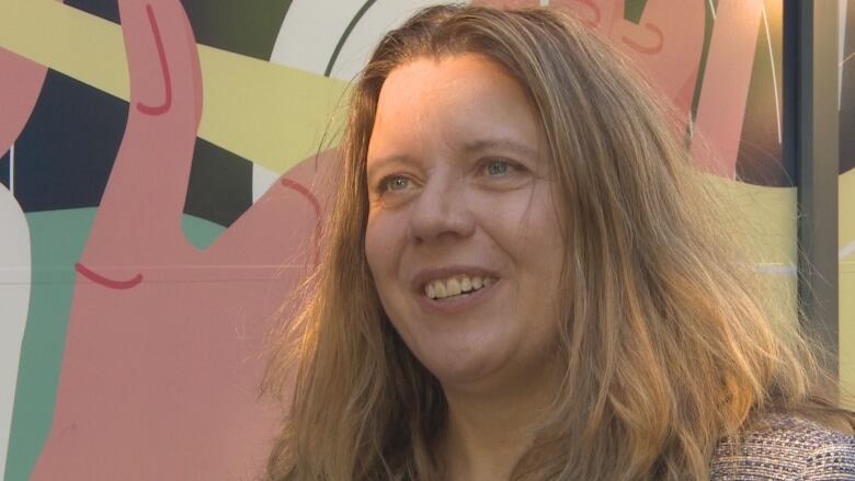 A close up of a woman  smiling as sun shines on her face and a mural in the background.