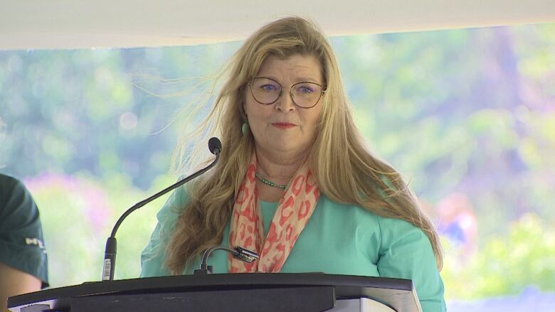 A woman with long blonde hair wearing a colorful scarf and a green cardigan speaks at a microphone during a news conference.