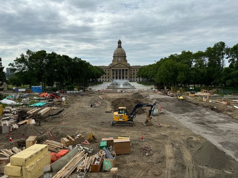 A construction site consisting of a dirt ground, piles of lumber, pipes and other materials is in the foreground with a yellow and black backhoe working on the site. The restored dome fountain and reflecting pool are in the distance, in front of the Alberta legislature building.