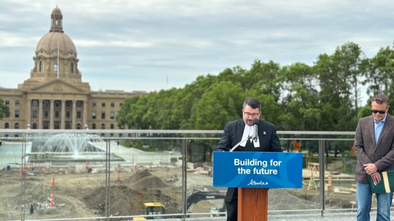 Speaker Nathan Cooper stands at a lectern at the centre of the photo, with Infrastructure Minister Pete Guthrie visible on his right. The lectern has a blue government sign that reads 