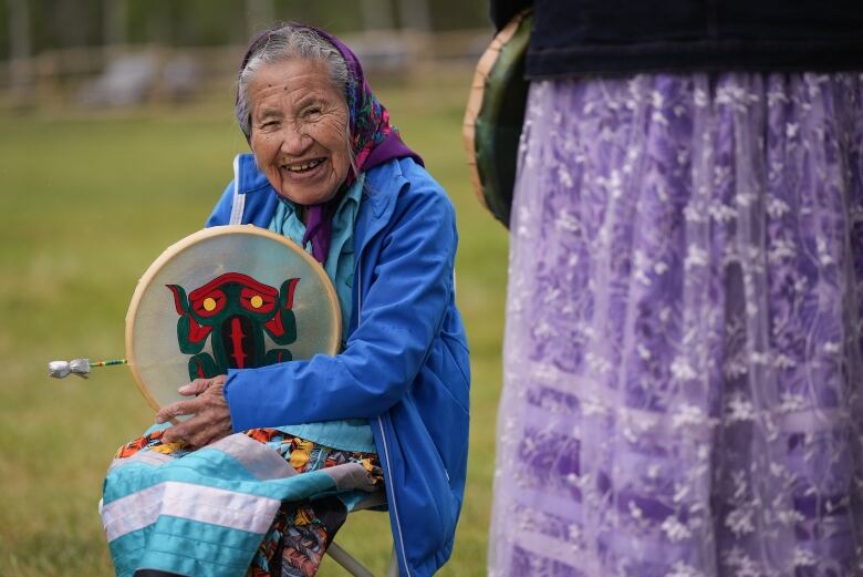 A woman holding a drum and wearing a blue jacket smiles for the camera while seated.