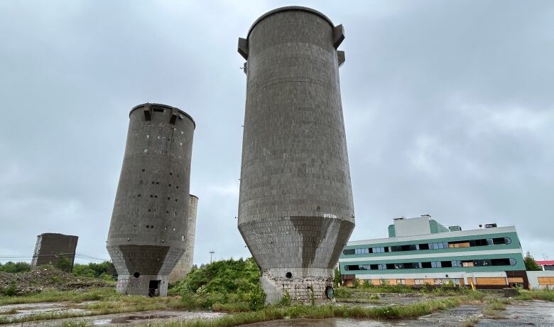 Three large cement cilos stand on an abandoned lot along with a green office building on a rainy day.