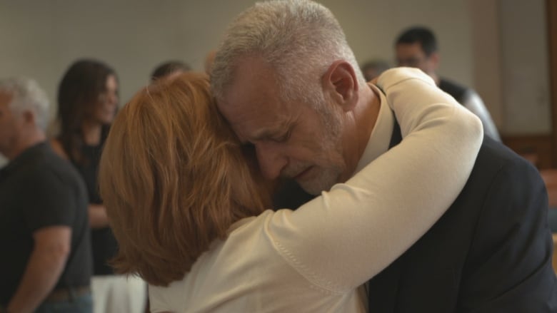 A man with grey hair and a beard is embraced by a woman in court.