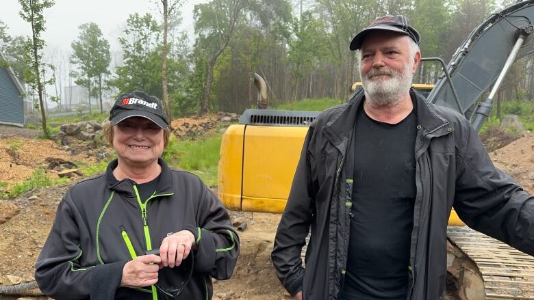 A man and a woman in black stand in front of an earthmover on a construction site.