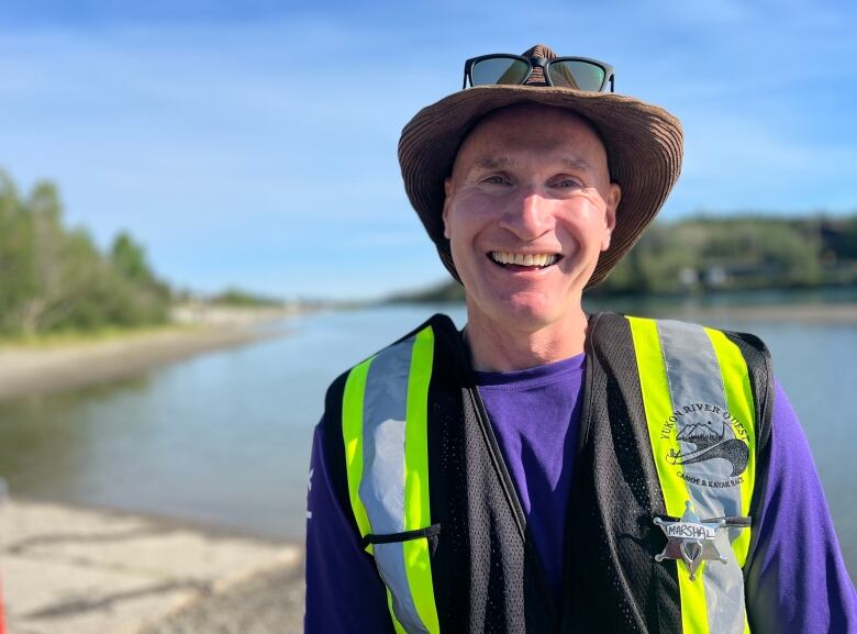 A man in a hat and safety vest stands beside a river on a sunny day.