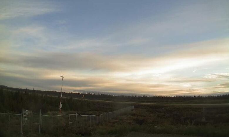 A webcam photo shows a pole against a field with a metal fence and cloudy sky.