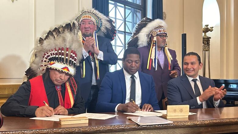 Men in First Nations traditional headdresses and two others in suits sign an agreement at a large table.